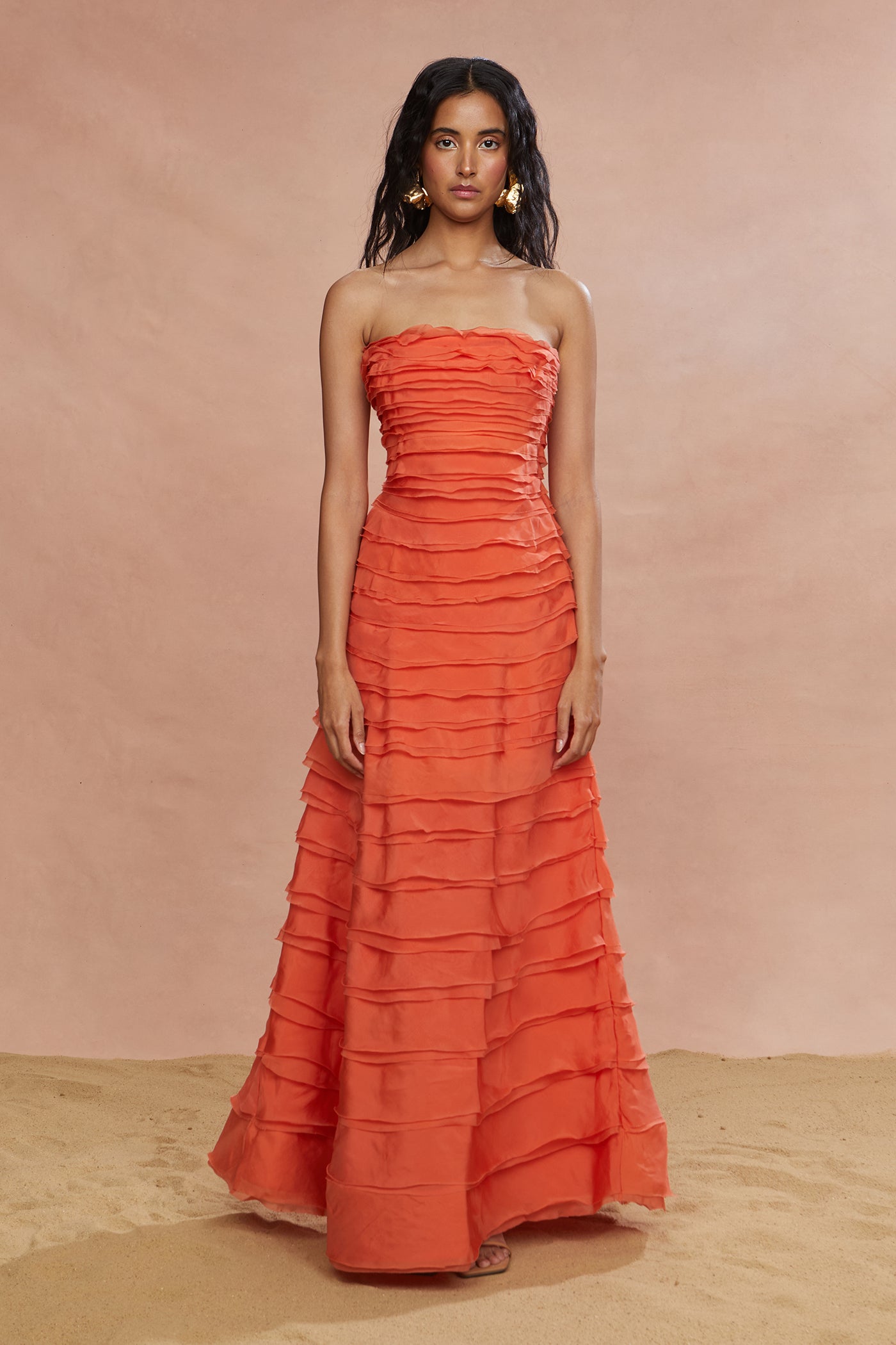 A woman standing in a studio setting wearing a strapless, orange, layered maxi dress.