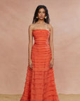 A woman standing in a studio setting wearing a strapless, orange, layered maxi dress.