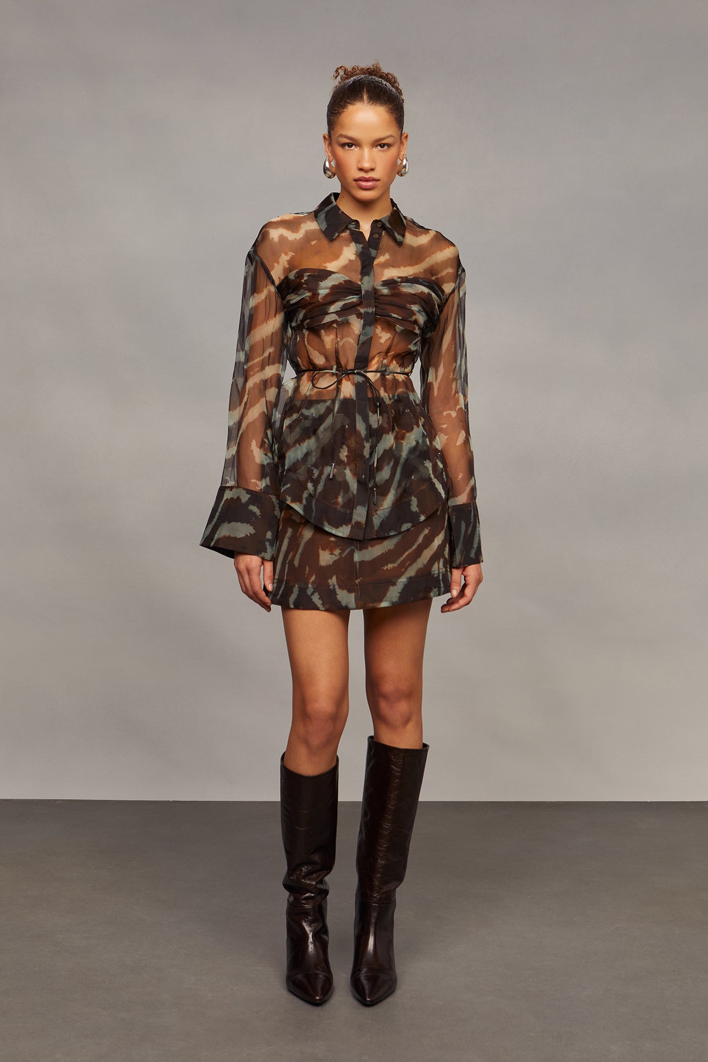 Model wearing a camouflage shirt with sheer sleeves, a matching skirt, and brown knee-high boots on a gray background