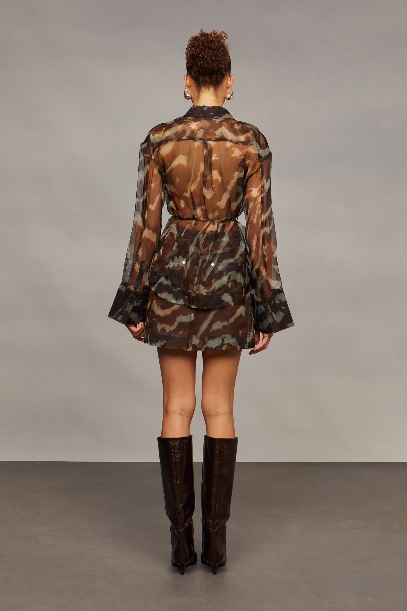 Model wearing a camouflage shirt with sheer sleeves, a matching skirt, and brown knee-high boots on a gray background