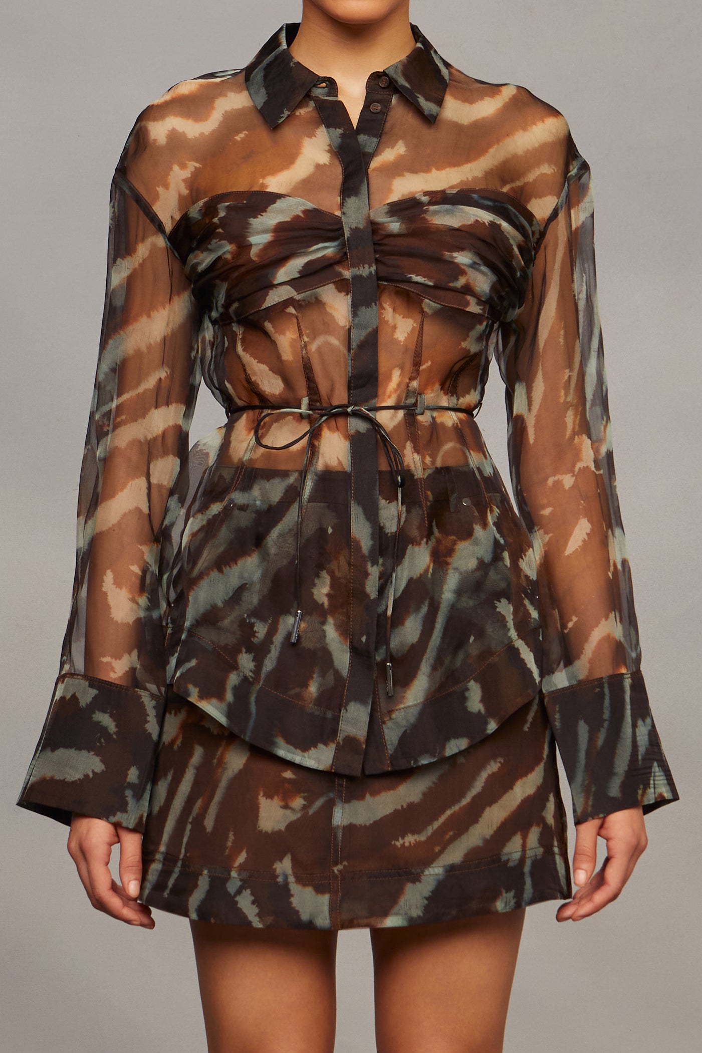 Model wearing a camouflage shirt with sheer sleeves, a matching skirt, and brown knee-high boots on a gray background