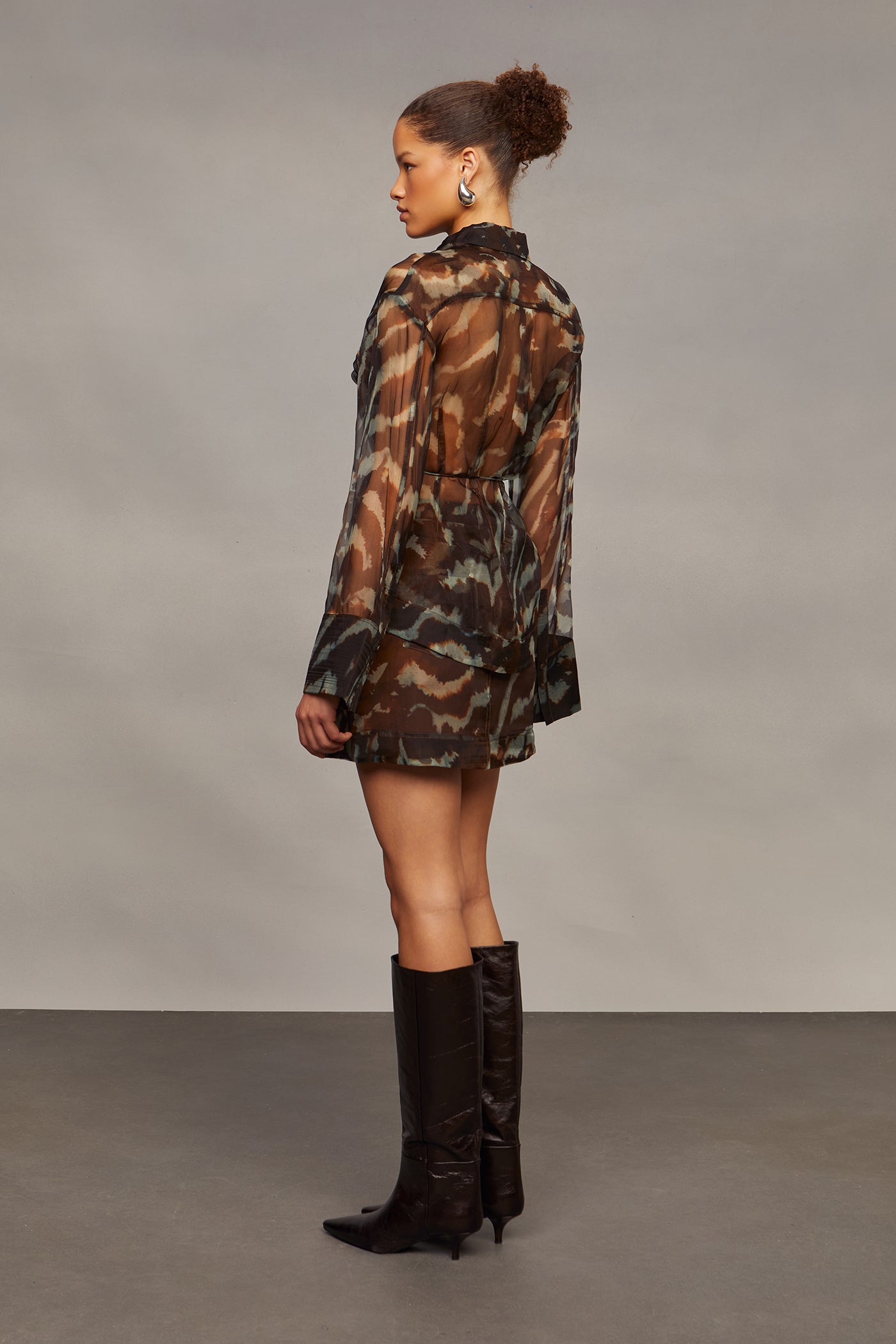 Model wearing a camouflage shirt with sheer sleeves, a matching skirt, and brown knee-high boots on a gray background