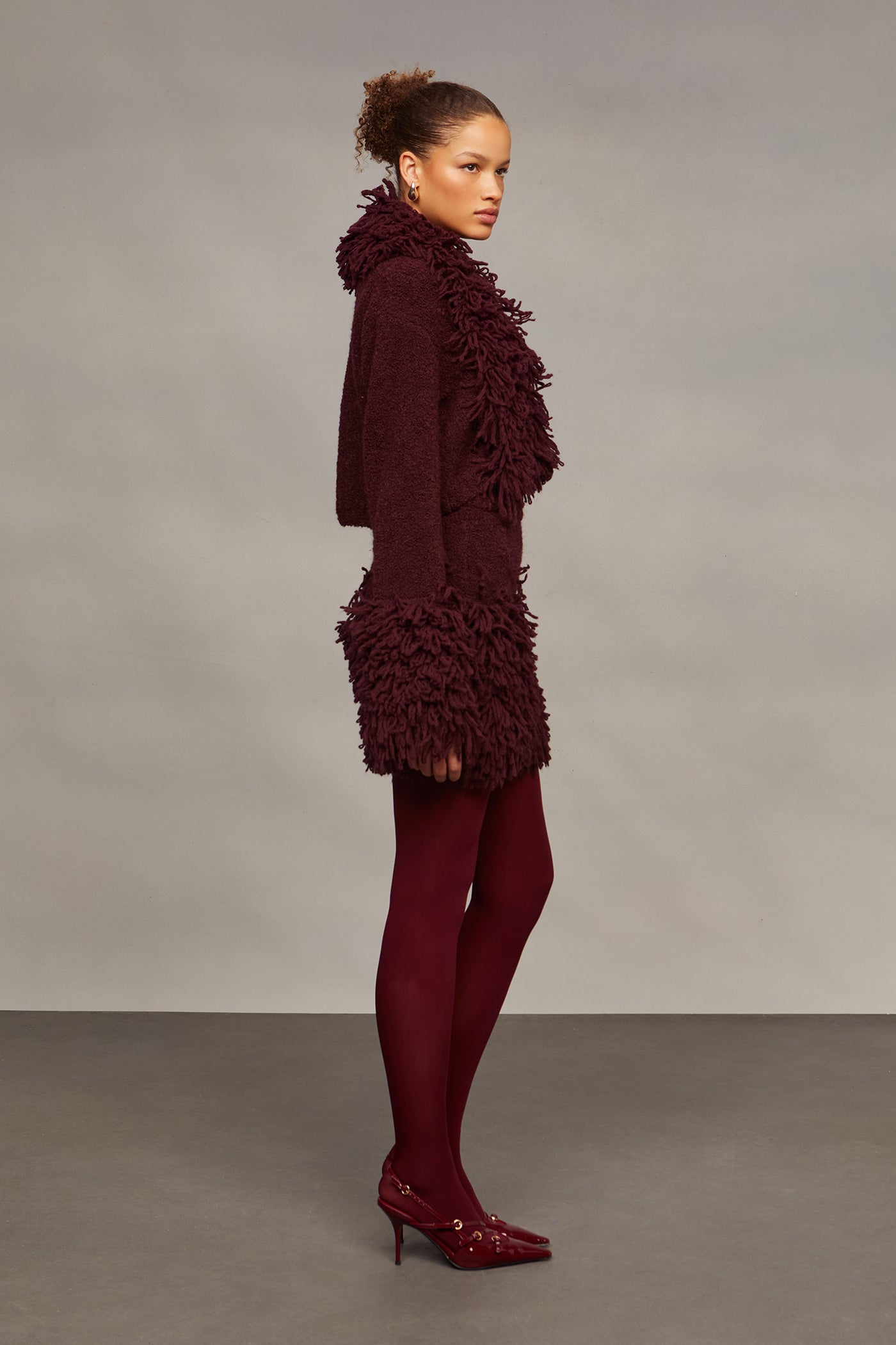 Woman wearing a burgundy textured outfit with matching tights and shoes against a gray background