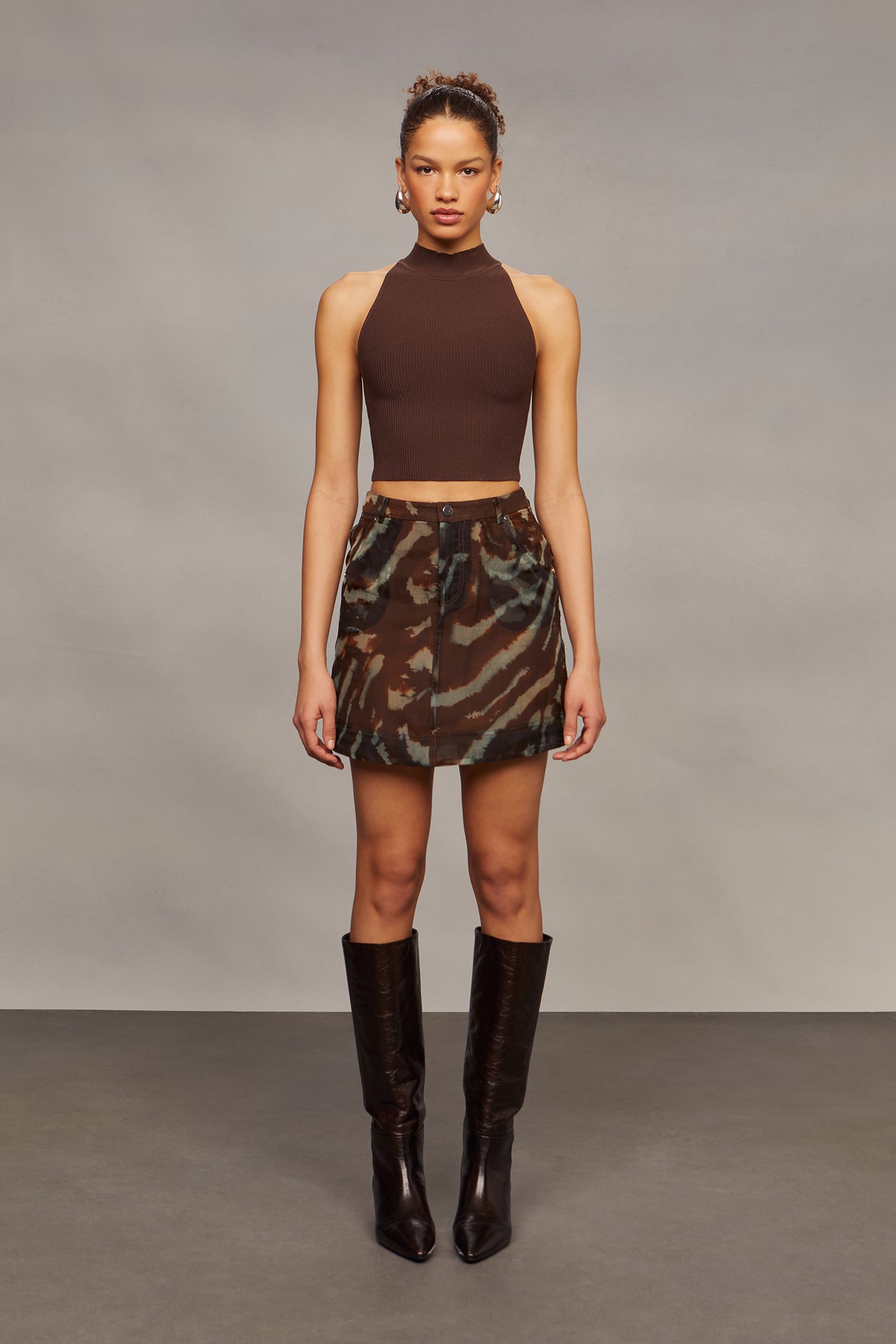 Woman wearing a brown halter top and camouflage skirt with brown knee-high boots against a gray background