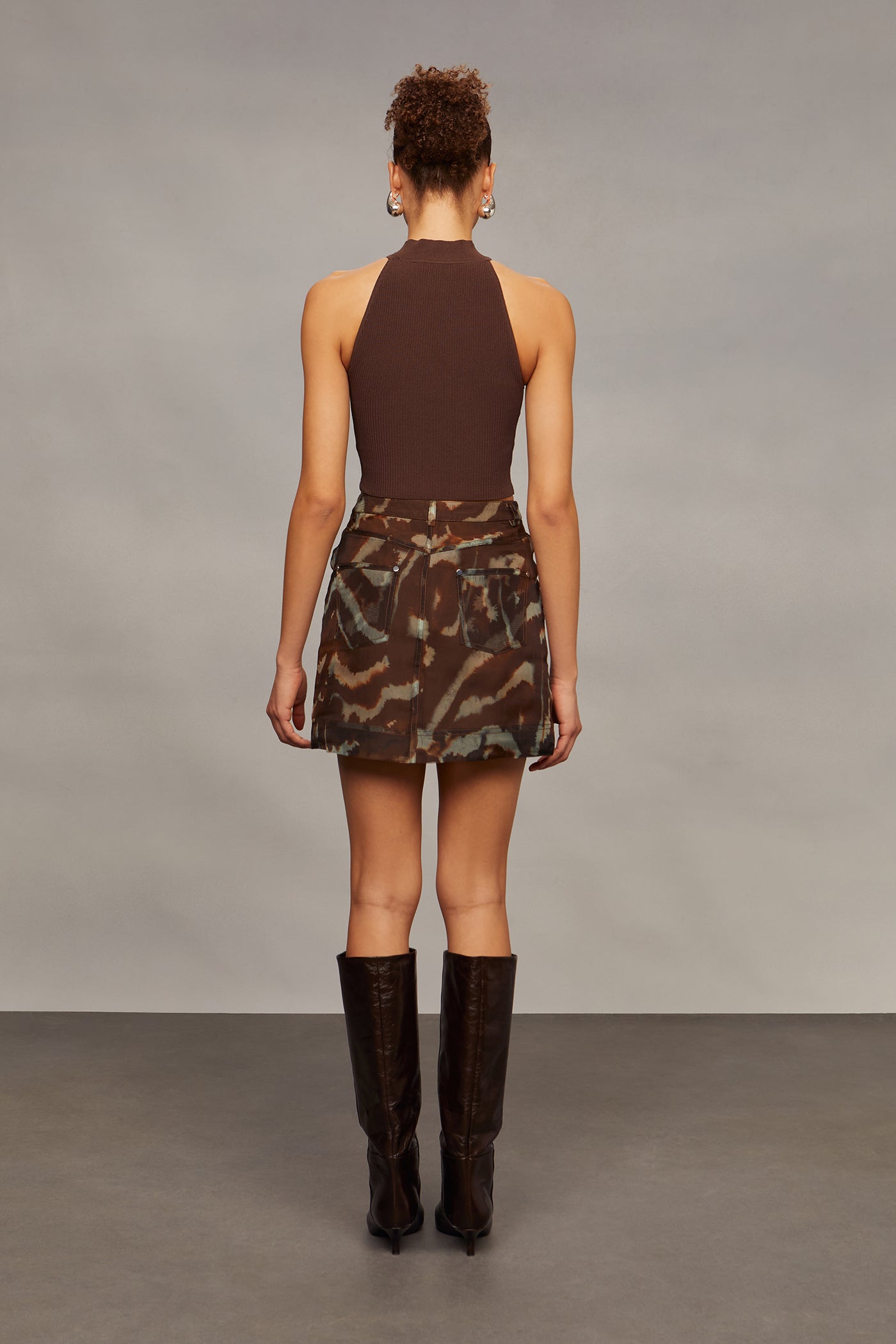 Woman wearing a brown halter top and camouflage skirt with brown knee-high boots against a gray background
