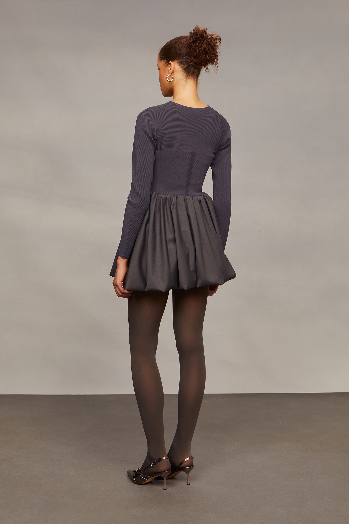 Person wearing a dark gray dress with long sleeves and a pleated skirt against a plain background